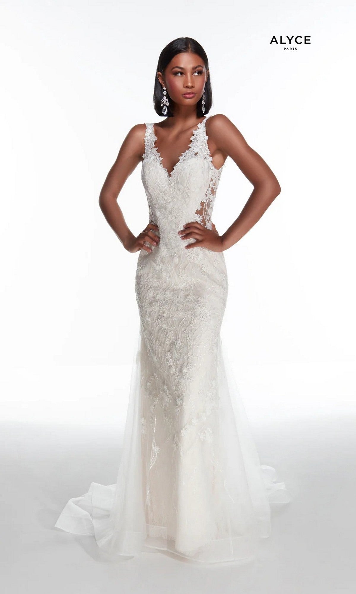 A woman poses in the Long White Dress: Alyce 7017, a sleeveless lace gown in Diamond White/Blush with a fitted bodice, sheer side panels, trumpet skirt, and long tulle train. The plain white background features the